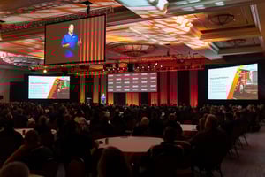 Conference general session featuring people sitting at round tables, four large screens, and speaker on stage.